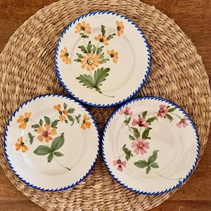 May include: Three white ceramic plates with hand-painted floral designs. Each plate features a different flower arrangement in shades of orange, pink, and green, with a blue zigzag border. The plates are arranged on a woven straw mat.