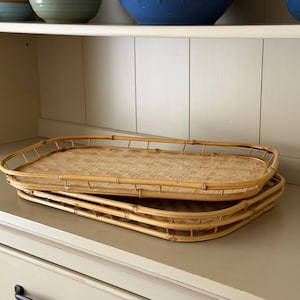May include: Two rectangular trays crafted from light brown natural rattan with a woven base and a raised border frame. The trays are stacked one on top of the other, featuring a consistent light tan color and a visible grid-like weave pattern.