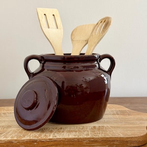 May include: A dark brown ceramic utensil holder with a lid, holding three wooden cooking utensils. The holder has two handles and sits on a wooden cutting board.