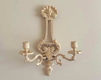 May include: A wall-mounted candle holder made of off-white painted metal with a distressed finish. The piece features a central vertical decorative element with scrollwork and two symmetrical arms, each supporting a single candle cup.