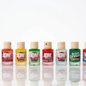 May include: Eight small car air fresheners in various colours, each with a wooden cap. Scents include espresso, sugar flower, lemon, and Irish Green. The bottles are arranged in a row, reflecting on a surface.