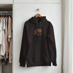 May include: A black hooded sweatshirt with a kangaroo pocket and a drawstring hood. The sweatshirt features a brown and tan illustration of a rabbit on the front. The hoodie is hanging on a wooden hanger.