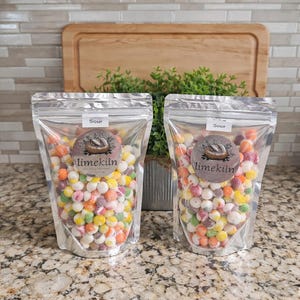 May include: Two clear, foil-lined bags filled with colorful, round candies. Each bag has a label that reads "Sour" and a circular sticker with the "limekiln" logo. The candies are a mix of orange, yellow, pink, green, and white.