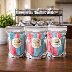 May include: Three clear plastic bags filled with red and blue candy sticks. Each bag has a round sticker with the text "limekiln" and a logo. The bags are labeled "Sour FR". The candy is arranged in vertical rows.