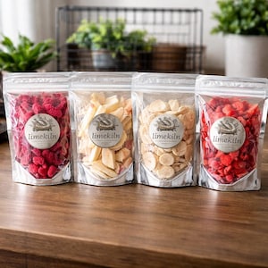 Puede incluir: Cuatro bolsas transparentes con base plateada llenas de fruta deshidratada. Contienen frambuesas rojas, chips de plátano claros y rodajas de almendras beige. Cada bolsa tiene una etiqueta circular con "limekiln". Sobre una superficie de madera.