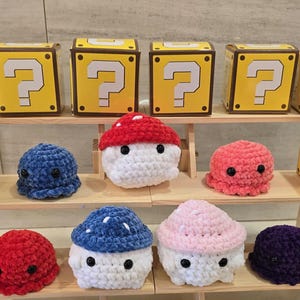 May include: A display of handmade crochet items, including mushroom-shaped figures, octopuses, and a frog, alongside yellow boxes with question mark designs. The items are arranged on a wooden tiered shelf.