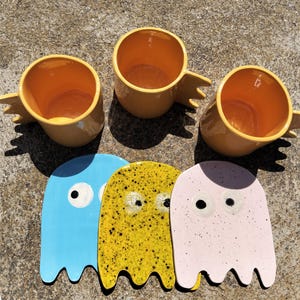 Può includere: Tre tazze in ceramica gialla con manici frastagliati disposte sopra tre sottobicchieri in ceramica a forma di fantasma. I sottobicchieri sono azzurri, gialli con macchie nere e rosa chiaro con macchie nere, ognuno con due occhi bianchi.