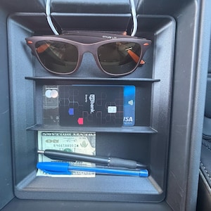 May include: A black plastic car center console organizer tray containing brown sunglasses, a black US Bank credit card, a blue Visa card, a twenty-dollar bill, a black ballpoint pen, and a blue ballpoint pen, all neatly arranged in separate compartments.
