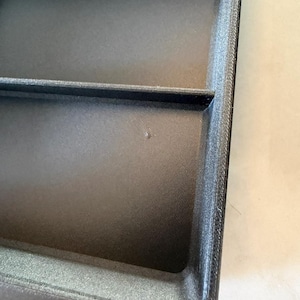 May include: A black rectangular storage tray featuring a smooth, matte finish and a single interior divider that separates the space into two distinct sections. The tray has raised edges and a textured surface with a small, singular mark.