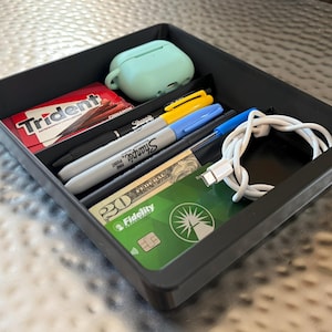 May include: A black plastic desk organizer tray containing a light green earbud case, a pack of Trident cinnamon gum, a yellow Sharpie, a blue Sharpie, a blue ballpoint pen, a white USB-C charging cable, a twenty-dollar bill, and a green Fidelity debit card.