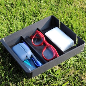 Peut inclure: Un plateau organisateur rectangulaire en plastique noir doté de trois compartiments distincts. Le plateau contient un étui à écouteurs bleu, un stylo bleu, une paire de lunettes à monture rouge et un petit récipient rectangulaire blanc.