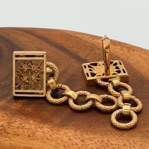 May include: Gold cufflinks with a rectangular face and a starburst design. The cufflinks are linked by textured, circular links. The background is a wooden surface. The cufflinks are made of gold.