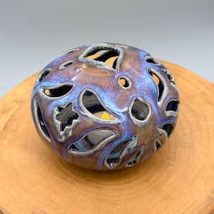 May include: A ceramic candle holder with a round shape and intricate cut-out designs. The candle holder has a blue and brown glaze, and is sitting on a wooden surface. The candle holder is designed to hold a small tea light.