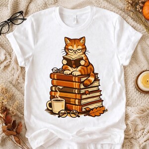 May include: White t-shirt featuring a cartoon cat wearing glasses and reading a book while sitting on a stack of books. A coffee mug, eyeglasses, and autumn leaves are also depicted in the design.