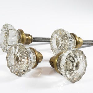 May include: Four clear glass door knobs with brass hardware. The knobs have a faceted, floral design. The hardware includes a metal rod for installation. The knobs are on a white background.