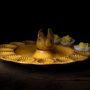 May include: A yellow ceramic deviled egg serving dish with a hen figurine in the center. The dish has a scalloped edge and spaces for eggs. Three deviled eggs are visible.