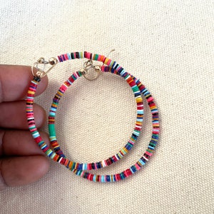 May include: A pair of gold hoop earrings adorned with colorful, small, flat beads. The beads are arranged in a pattern of red, pink, blue, green, and yellow. The earrings are displayed against a neutral background.