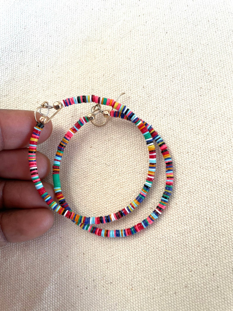 May include: A pair of gold hoop earrings adorned with colorful, small, flat beads. The beads are arranged in a pattern of red, pink, blue, green, and yellow. The earrings are displayed against a neutral background.