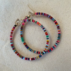 May include: A pair of colorful hoop earrings. The earrings are made of small, flat, round beads in a variety of colors, including pink, red, blue, green, and yellow. The earrings have silver-colored ear wires.
