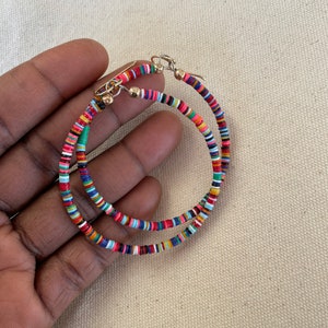 May include: A pair of hoop earrings with a colorful, beaded design. The earrings feature small, flat, round beads in various colors, including red, blue, green, and yellow. The hoops are held together with gold-colored hooks.