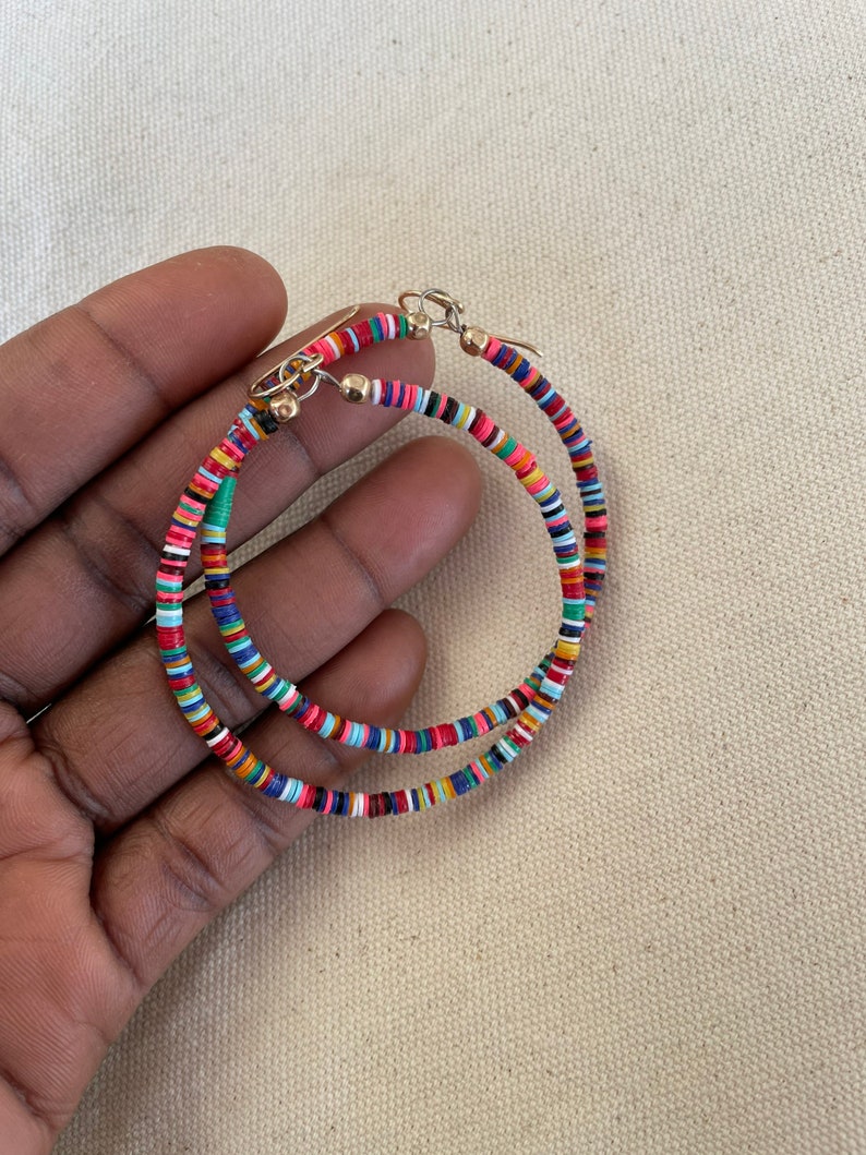 May include: A pair of hoop earrings with a colorful, beaded design. The earrings feature small, flat, round beads in various colors, including red, blue, green, and yellow. The hoops are held together with gold-colored hooks.