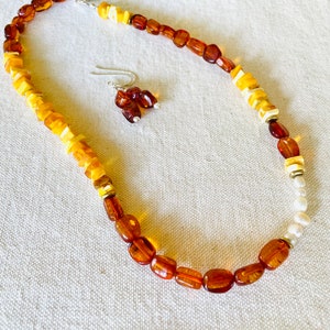 May include: A necklace and earrings set featuring amber and white shell beads. The necklace is graduated in color, with darker amber beads at the bottom and lighter amber beads at the top. The earrings are simple and feature two amber beads each.