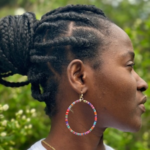 May include: Large hoop earrings with a gold-colored hook. The hoops are adorned with small, colorful beads in shades of red, orange, blue, green, and purple. The earrings are worn by a person with dark skin and braided hair.