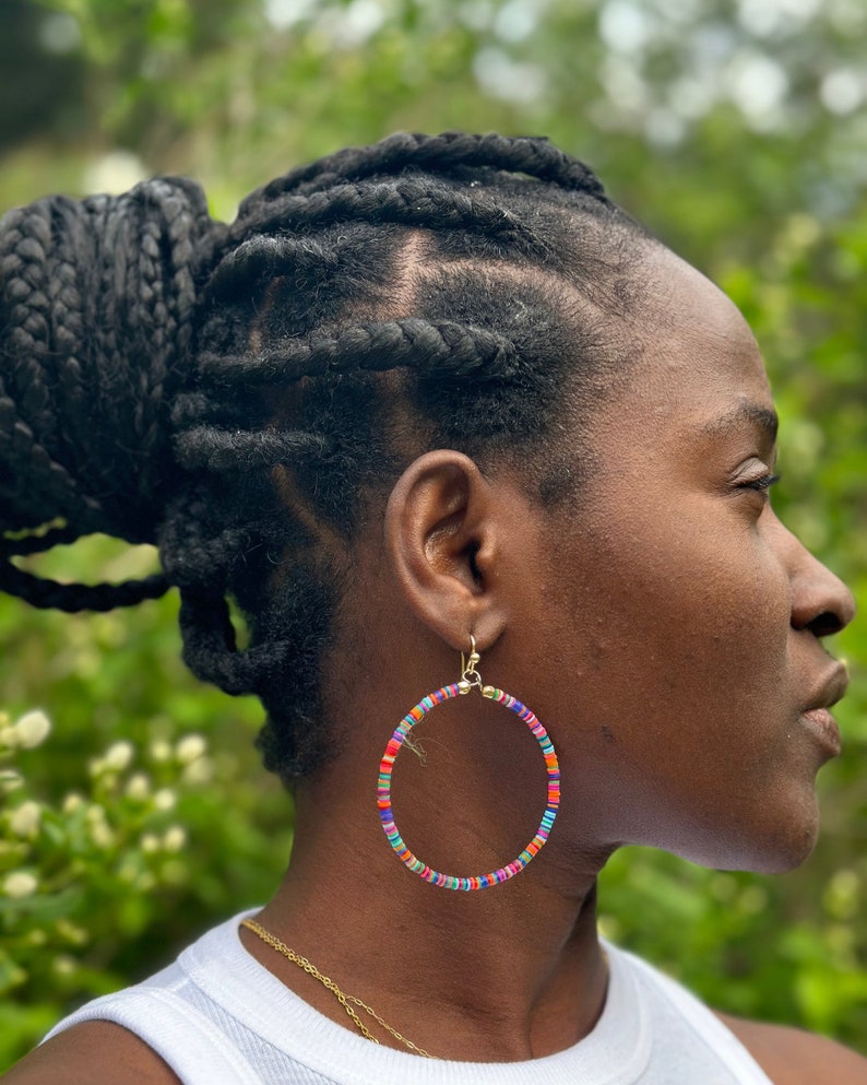 May include: Large hoop earrings with a gold-colored hook. The hoops are adorned with small, colorful beads in shades of red, orange, blue, green, and purple. The earrings are worn by a person with dark skin and braided hair.