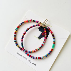 May include: Two colorful beaded hoop earrings with gold-colored clasps. The earrings are made of small, flat, round beads in various colors, including red, pink, yellow, green, and blue. The earrings are displayed on a white card with the website address www.akweleydesign.com.