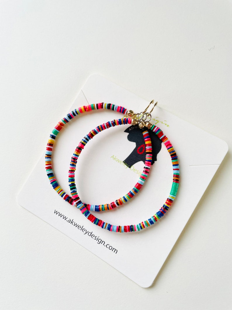 May include: Two colorful beaded hoop earrings with gold-colored clasps. The earrings are made of small, flat, round beads in various colors, including red, pink, yellow, green, and blue. The earrings are displayed on a white card with the website address www.akweleydesign.com.