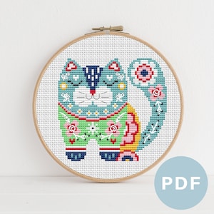 May include: Cross stitch pattern of a colorful cat with floral details. The cat is wearing a blue and green outfit with yellow and pink accents. The pattern is in a round embroidery hoop.
