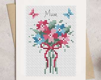 May include: A white card with a cross-stitch design of a bouquet of flowers in shades of pink, blue, and green. The flowers are tied with a red ribbon and there are two butterflies in the top corners. The card says "Mum" at the top.
