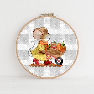 May include: Cross-stitch embroidery in a wooden hoop depicts a mouse in a yellow coat and red boots pushing a wheelbarrow filled with pumpkins and apples. The mouse is wearing an orange scarf. The background is white.