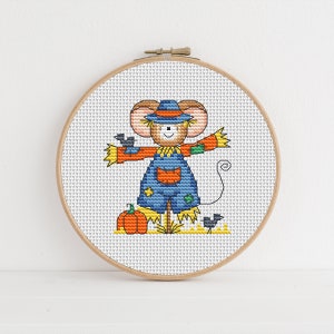 May include: A cross-stitch pattern of a cute mouse dressed as a scarecrow with a pumpkin and birds. The mouse is wearing a blue and orange outfit with a straw hat.