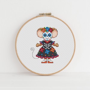 May include: Cross-stitch embroidery of a mouse dressed in a black dress with colorful floral and skeleton details. The mouse has a floral crown and is set in a wooden embroidery hoop. The design is a Day of the Dead theme.