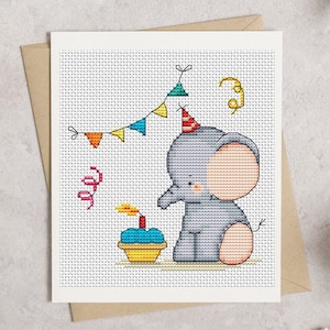May include: A cross-stitch pattern on a white card featuring a grey elephant wearing a red and white striped party hat. The elephant sits next to a blue cupcake with a lit candle. Above are colorful triangular bunting flags and streamers.