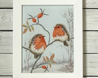 May include: A framed cross-stitch embroidery featuring two robins with orange breasts perched on thin branches. The birds are set against a light blue background with small orange berries and green leaves. The piece is enclosed in a simple white frame.