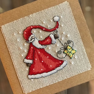 Può includere: Un biglietto di punto croce con un Babbo Natale rosso e bianco che tiene un regalo giallo e verde. Lo sfondo è un tessuto di lino beige con fiocchi di neve bianchi.