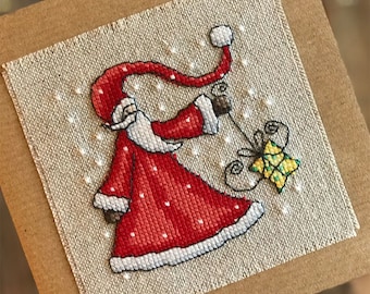May include: A cross-stitch card featuring a red and white Santa Claus holding a yellow and green gift. The background is a beige linen fabric with white snowflakes.