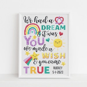 May include: A framed cross-stitch design with a rainbow, stars, and a sun. The text reads "We had a dream & it was you. We made a wish & you came true. Aubrey 5-1-2023".
