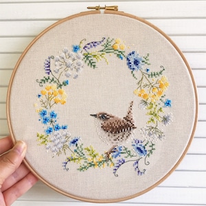 May include: A brown and white bird with a long beak sits in a wreath of yellow, blue, and white flowers. The wreath is embroidered on a beige fabric and is framed in a wooden hoop.