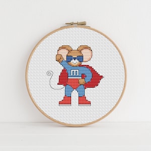 May include: A cross-stitch embroidery hoop featuring a design of a brown mouse dressed as a superhero, wearing a red and blue costume with a red cape and the letter "M" on the chest.