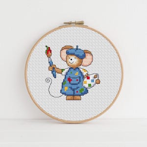 May include: A circular wooden embroidery hoop containing a cross-stitch pattern of a brown mouse wearing a blue beret and blue overalls. The mouse holds a paintbrush in one hand and a white paint palette with colorful paint spots in the other.