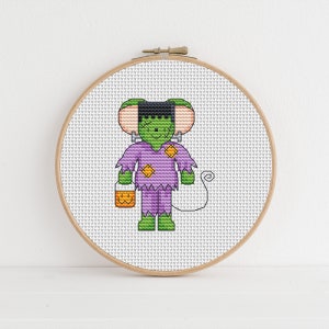 May include: Cross stitch pattern of a green monster wearing a purple costume and holding a pumpkin bucket. The pattern is in a wooden embroidery hoop.