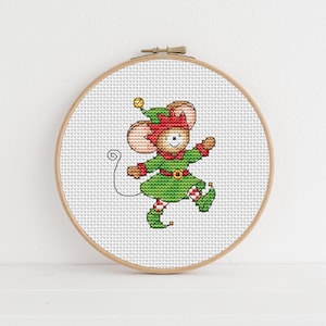 May include: Cross-stitch embroidery of a mouse dressed as an elf, set in a wooden hoop. The mouse wears a green outfit with red accents, a matching hat, and a gold belt buckle. The background is white, and the design is centered.