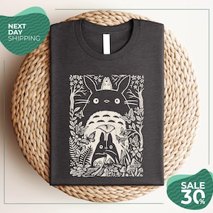 a gray t-shirt with a black and white graphic design depicting a cat surrounded by various natural elements, such as flowers, leaves, and mushrooms. The t-shirt is placed on a woven placemat, and there are two green circular signs in the image. One sign advertises "Next Day Shipping" and the other sign advertises a "SALE 30%".