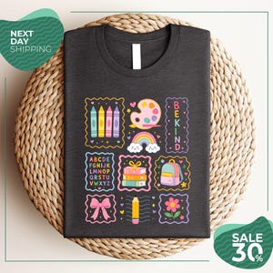 a black t-shirt with a colorful and whimsical design, including various school supplies and educational symbols such as crayons, a paint palette, a backpack, and a rainbow. The t-shirt is displayed on a woven basket, and there are two green circular signs in the image, one on the left side and one on the right side, which appear to be related to shipping or a sale.