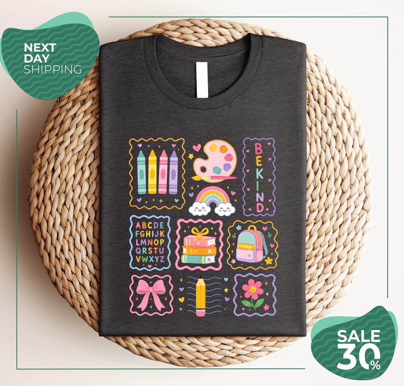 a black t-shirt with a colorful and whimsical design, including various school supplies and educational symbols such as crayons, a paint palette, a backpack, and a rainbow. The t-shirt is displayed on a woven basket, and there are two green circular signs in the image, one on the left side and one on the right side, which appear to be related to shipping or a sale.