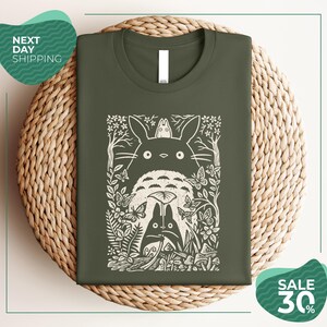 a green t-shirt with a white graphic design depicting a cat surrounded by various natural elements, such as leaves, flowers, and mushrooms. The t-shirt is displayed on a woven placemat, and there are two green circular signs in the image, one indicating "Next Day Shipping" and the other promoting a "SALE 30%".