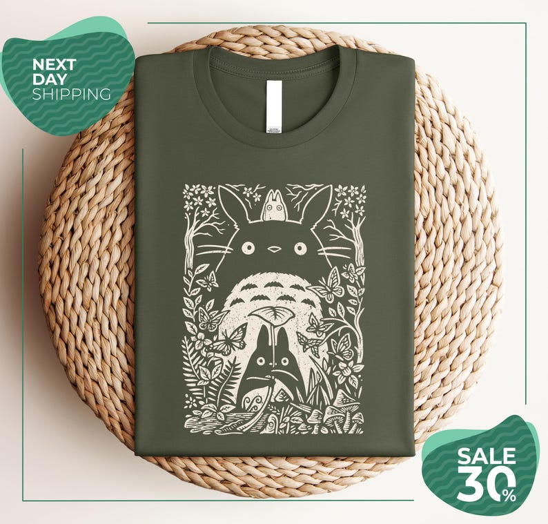 a green t-shirt with a white graphic design depicting a cat surrounded by various natural elements, such as leaves, flowers, and mushrooms. The t-shirt is displayed on a woven placemat, and there are two green circular signs in the image, one indicating "Next Day Shipping" and the other promoting a "SALE 30%".